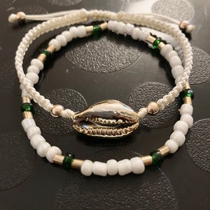 May include: Two white beaded bracelets with gold and green accents. The bracelets are tied together with a white cord and feature a gold seashell charm.
