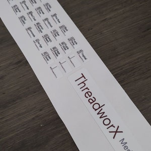 Puede incluir: Una hoja de etiquetas blancas con texto negro, que presenta una variedad de colores y nombres de hilos. Las etiquetas est&aacute;n dispuestas en filas y columnas. La parte inferior de la hoja presenta un logotipo rojo y blanco que dice "ThreadworX Merino".