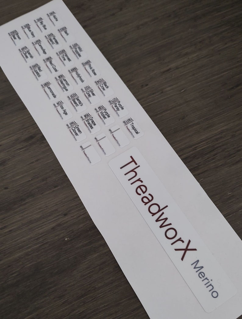 Puede incluir: Una hoja de etiquetas blancas con texto negro, que presenta una variedad de colores y nombres de hilos. Las etiquetas est&aacute;n dispuestas en filas y columnas. La parte inferior de la hoja presenta un logotipo rojo y blanco que dice "ThreadworX Merino".