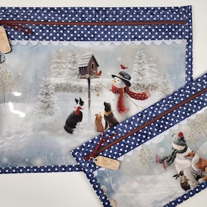 Puede incluir: Dos estuches azules con cremallera y ventanas de vinilo transparente. El estuche más grande presenta una escena de invierno con un muñeco de nieve, una casita para pájaros y perros. El estuche más pequeño presenta un muñeco de nieve con un zorro y un conejo. Ambos estuches tienen una cremallera roja y un patrón de lunares blancos.