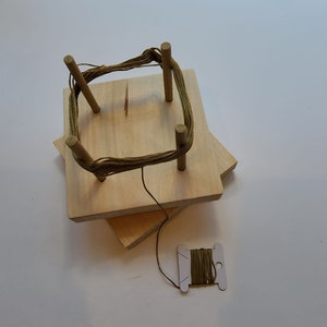 Op de afbeelding: Een houten garenwinder met vier pinnen en een spoel met groene draad. De winder is gemaakt van twee stukken hout die op elkaar zijn gestapeld.