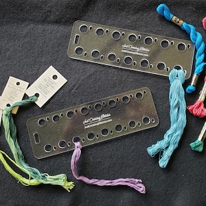 May include: Two clear plastic embroidery floss organizers with multiple holes for storing floss.  The organizers are labeled "Just Cross Stitch" and "JustCrossStitching.com".  Several skeins of embroidery floss in various colors are shown next to the organizers.