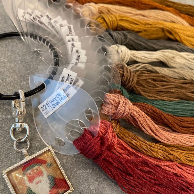 Puede incluir: Primer plano de un organizador de hilo de bordar de pl&aacute;stico con una variedad de colores de hilo enrollados alrededor. El organizador tiene un anillo de pl&aacute;stico transparente y un cierre de metal. Los colores del hilo incluyen rojo, naranja, amarillo, verde, marr&oacute;n y rosa. El organizador tambi&eacute;n tiene un peque&ntilde;o amuleto adjunto con una imagen de Pap&aacute; Noel.