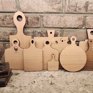 May include: A collection of 10 wooden chopping boards of various shapes and sizes. The boards are made of light brown wood and have a natural finish. Some of the boards have a small handle on the top. The boards are arranged in a row against a brick wall background.