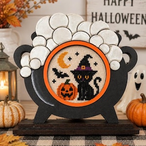 May include: A black wooden cauldron-shaped frame with a bright orange circular border containing a cross-stitch design of a black cat wearing a purple hat, a yellow crescent moon, a bat, and a jack-o'-lantern, topped with white wooden bubbles.