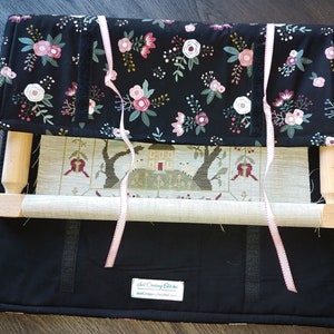 May include: A wooden embroidery hoop with a floral fabric cover and a pink ribbon. The hoop is on a black fabric background with a label that reads "Just Crafty Stitches".