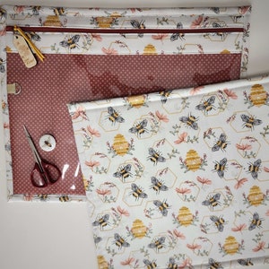 Könnte beinhalten: Eine rote und weiße Stofftasche mit Reißverschluss und einem transparenten Plastikfenster. Die Tasche ist mit einem sich wiederholenden Muster aus Waben und Bienen verziert. Die Tasche liegt auf einer weißen Oberfläche mit einem weißen Stoff mit einem ähnlichen Muster.