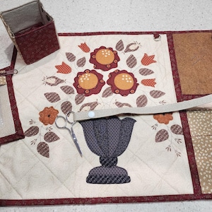 May include: A quilted sewing organizer with a floral design, featuring a vase with flowers in shades of orange, red, and brown. Includes a zippered pouch with sewing tools, a small box, and two brown fabric pieces. A pair of scissors is also visible.