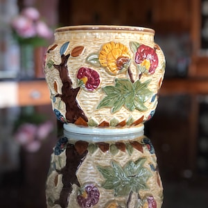 May include: A ceramic planter with a raised floral design. The planter is cream coloured with brown, green, yellow, and red flowers. The planter is sitting on a black surface.