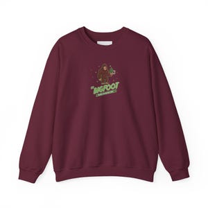 Puede incluir: Sudadera burdeos con un gráfico de Bigfoot. El gráfico presenta un Bigfoot de dibujos animados sosteniendo un objeto verde, con la palabra "BIGFOOT" en texto verde. Debajo del texto está "Pacific Northwest, USA".