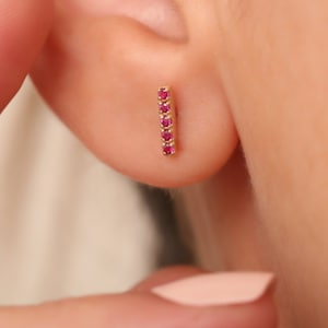 Op de afbeelding: Een gouden oorbel in de vorm van een staafje met vijf kleine, helderrode edelstenen in een verticale lijn. De oorbel wordt op een oor getoond, wat het ontwerp en het kleurcontrast benadrukt. Het sieraad is eenvoudig maar elegant.