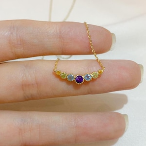 Op de afbeelding: Een gouden ketting met een klein hangertje met vijf gekleurde edelstenen in een gebogen vorm. De edelstenen zijn geel, blauw, paars, blauw en geel.