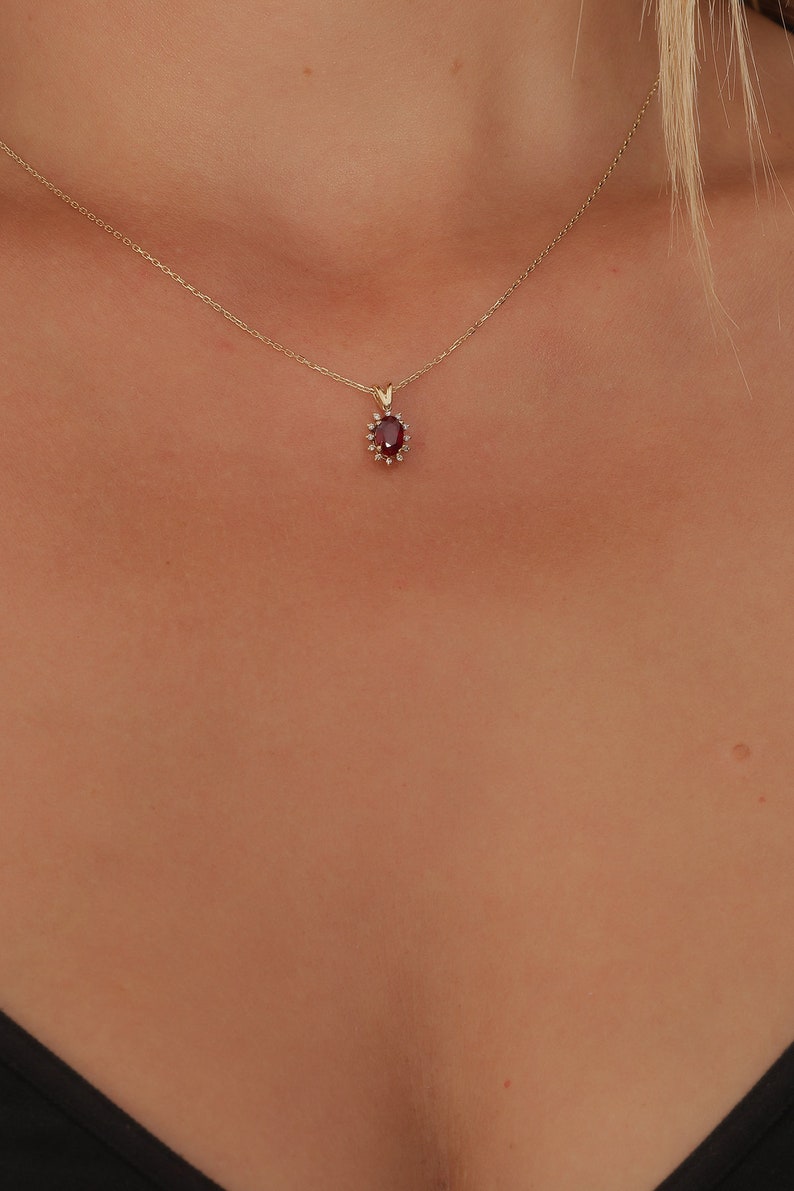 Op de afbeelding: Een gouden ketting met een rode edelsteen hanger omringd door kleinere heldere edelstenen.