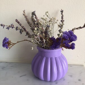 May include: A small, round, lavender-colored vase with a ribbed design, filled with dried lavender and other purple and white flowers. The vase sits on a white marble surface.