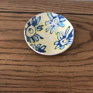 Peut inclure: Un petit plat en céramique blanche avec un motif floral bleu et jaune. Le plat a une forme arrondie et un bord légèrement surélevé.