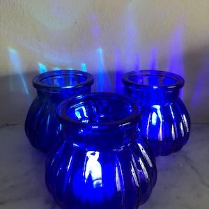 May include: Three cobalt blue glass candle holders with a ribbed design. The candle holders are lit and glowing.