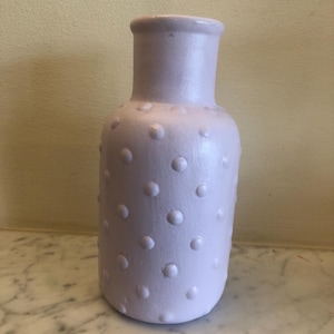 May include: A lilac-coloured vase with a textured surface of raised dots. The vase has a cylindrical neck and a rounded body. The vase is made of ceramic or a similar material and is suitable for displaying flowers or as a decorative item.