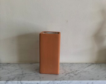 May include: A terracotta colored ceramic vase with a square shape. The vase is sitting on a white marble surface.