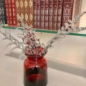 May include: A red glass vase with a silver fern and red berries on a white shelf with a row of books behind it. The books are all bound in red leather with gold lettering.