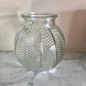 May include: A clear glass vase with a spherical body and a textured leaf pattern. The vase has a wide opening. It is displayed on a marble surface. The design gives the vase a vintage or botanical aesthetic.
