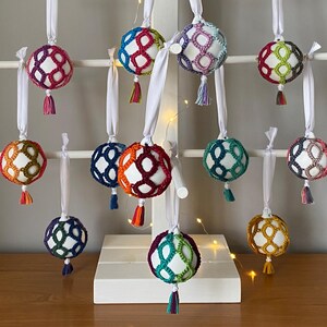 May include: A collection of colourful, crocheted baubles with white ribbon hangers and small tassels. The baubles are in various colours, including red, blue, green, and orange, and are displayed on a white wooden tree.