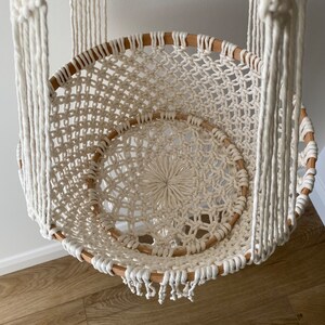 May include: A cream-coloured macrame hanging chair with a circular design. The chair is made of woven rope and features a wooden frame. The seat has a woven pattern, and the chair hangs from thick, braided ropes.