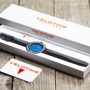 Op de afbeelding: Een witte doos met de tekst "VELOTIME WATCHES" in oranje gedrukt. De doos bevat een zwart horloge met een blauwe wijzerplaat en oranje accenten. Het horloge is aan een zwart leren bandje.