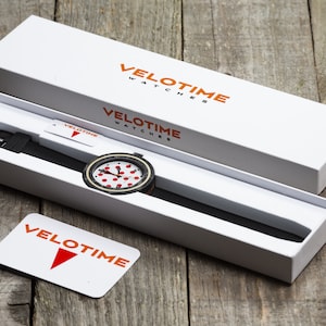 Op de afbeelding: Een witte horloge doos met een zwart-wit horloge met rode stippen erin. De doos heeft de tekst "VELOTIME WATCHES" erop gedrukt in oranje letters. Een kleine witte kaart met de tekst "VELOTIME" gedrukt in rode letters is ook zichtbaar.