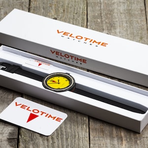 Op de afbeelding: Een witte doos met de tekst "VELOTIME WATCHES" op de bovenkant en een geel en zwart horloge met een zwarte band erin. De wijzerplaat van het horloge heeft een rood en wit ontwerp.