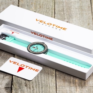 Op de afbeelding: Een witte doos met de tekst "VELOTIME WATCHES" op de bovenkant en "VELOTIME" aan de zijkant. De doos is open en er zit een horloge met een groene band in. Het horloge heeft een zwarte wijzerplaat met witte cijfers en een houten bezel.