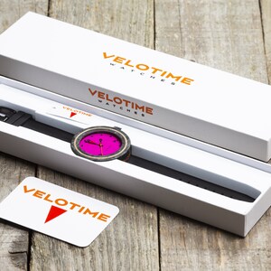 Op de afbeelding: Een zwart en roze polshorloge met een houten wijzerplaat en een zwarte leren band wordt getoond in een witte doos met het Velotime Watches logo. De doos is open en het horloge is gedeeltelijk zichtbaar.