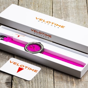 Op de afbeelding: Een witte doos met de tekst "VELOTIME WATCHES" gedrukt op de bovenkant en zijkant. De doos is open en er zit een roze horloge met een zwarte wijzerplaat in. Het horloge heeft een zwarte band en een witte wijzerplaat met zwarte cijfers. Het horloge ligt op een houten oppervlak.