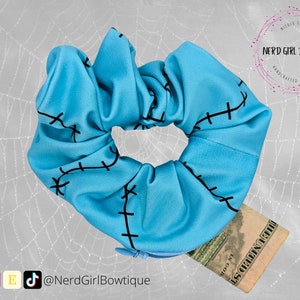 Puede incluir: Un scrunchie azul con costuras negras, que se asemeja a una herida cosida. El scrunchie está cubriendo parcialmente un billete de dólar.