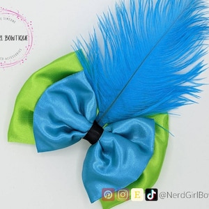 May include: A blue and green satin bow with a blue feather on top. The bow is handmade and has a black center. The feather is long and fluffy. The bow is perfect for adding a touch of whimsy to any outfit.