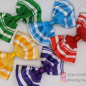 May include: Six striped fabric hair bows in different colors. The bows are arranged in a circle on a white background. The colors of the bows are green, blue, yellow, red, purple, and light blue. The bows are all made of the same fabric, but they have different colored stripes. The bows are all tied with a white ribbon.