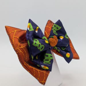 Puede incluir: Un lazo para el pelo con tem&aacute;tica de Halloween. Cuenta con un lazo azul oscuro con la palabra "Boo" en verde, caramelos de ma&iacute;z y una peque&ntilde;a calabaza naranja, sobre una base naranja con dise&ntilde;o de telara&ntilde;a.