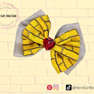 Puede incluir: Un lazo para el cabello con tela amarilla con un patrón de ladrillos negros sobre una tela gris claro lisa. El centro del lazo está adornado con una gema roja facetada en forma de corazón. El texto Nerd Girl Bowtique es visible en la esquina.