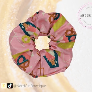 May include: Pink scrunchie with a repeating pattern of ice cream cones, pretzels, and geometric shapes. The scrunchie is a hair accessory and features the text "Nerd Girl Boutiquie".
