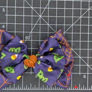Puede incluir: Un lazo para el pelo con tem&aacute;tica de Halloween, con tela morada oscura con un dise&ntilde;o de telara&ntilde;a naranja. El lazo central presenta caramelos de ma&iacute;z y las letras "Dw" y "ma". Un centro de calabaza con purpurina naranja.