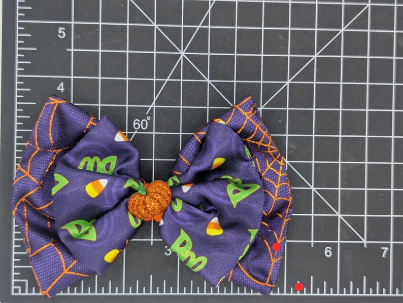 Puede incluir: Un lazo para el pelo con tem&aacute;tica de Halloween, con tela morada oscura con un dise&ntilde;o de telara&ntilde;a naranja. El lazo central presenta caramelos de ma&iacute;z y las letras "Dw" y "ma". Un centro de calabaza con purpurina naranja.