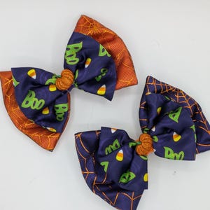 Puede incluir: Tres lazos para el pelo con tem&aacute;tica de Halloween. Los lazos presentan una tela azul marino con texto verde "Boo" y detalles de caramelos de ma&iacute;z. Los bordes son naranjas con un dise&ntilde;o de telara&ntilde;a. Cada lazo tiene un centro naranja brillante.