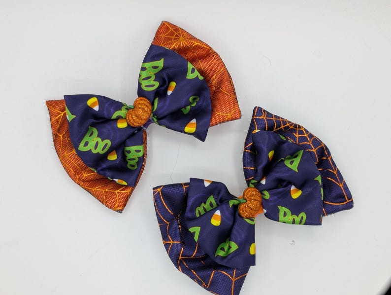 Puede incluir: Tres lazos para el pelo con tem&aacute;tica de Halloween. Los lazos presentan una tela azul marino con texto verde "Boo" y detalles de caramelos de ma&iacute;z. Los bordes son naranjas con un dise&ntilde;o de telara&ntilde;a. Cada lazo tiene un centro naranja brillante.
