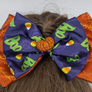 Puede incluir: Un lazo para el pelo con tem&aacute;tica de Halloween con un centro morado con patrones de "Boo" y caramelos de ma&iacute;z. El lazo tiene lados naranjas con un dise&ntilde;o de telara&ntilde;a y un centro de calabaza brillante. Un accesorio festivo para Halloween.
