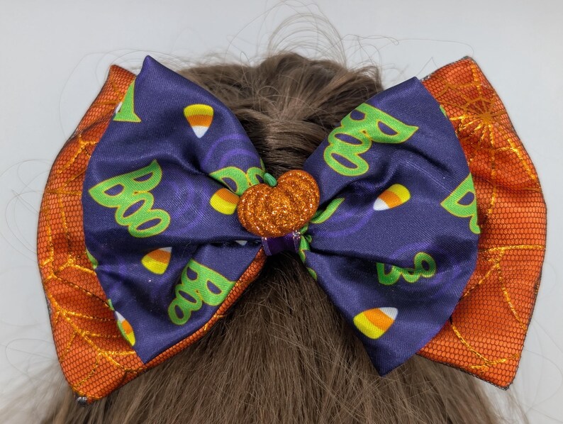 Puede incluir: Un lazo para el pelo con tem&aacute;tica de Halloween con un centro morado con patrones de "Boo" y caramelos de ma&iacute;z. El lazo tiene lados naranjas con un dise&ntilde;o de telara&ntilde;a y un centro de calabaza brillante. Un accesorio festivo para Halloween.