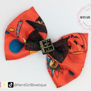 May include: A red, orange, and black bow with a witch hat, pumpkin, and "Book of Spells" design. The bow is made of satin fabric and has a black mesh overlay.