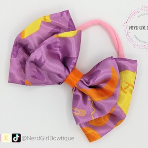 May include: Purple and orange satin bow with a pink elastic headband. The bow has a pattern of yellow moons and orange light bulbs. The headband is for hair.
