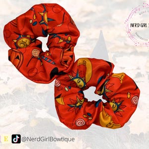 May include: Two red scrunchies with a celestial print. The print features suns, moons, and stars in yellow, orange, and blue. The scrunchies are made of satin.