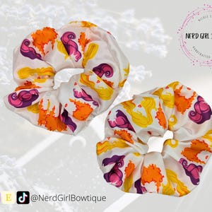 May include: Two white satin scrunchies with a yellow, orange, and purple floral print. The scrunchies feature a cartoon-like design of a woman with long blonde hair.