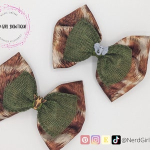 May include: Two handcrafted hair bows with a safari theme. The bows feature a brown and tan animal print fabric, olive green burlap centers, and small animal embellishments. The Nerd Girl Bowtique logo is in the upper left corner.