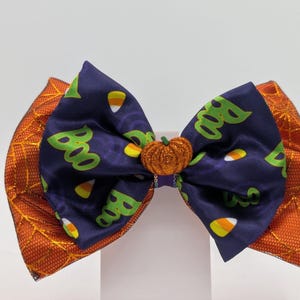 Puede incluir: Un lazo para el pelo con tem&aacute;tica de Halloween con un centro azul marino con patrones de "Boo" y caramelos de ma&iacute;z. Tiene detalles laterales naranjas y un adorno de calabaza brillante. Un accesorio festivo para Halloween.